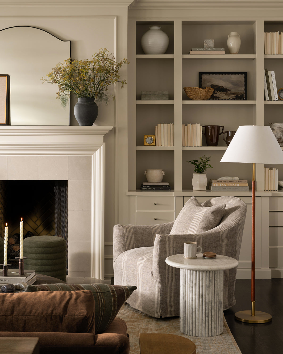 A cozy living room features a light armchair by a round marble side table and the Elijah Floor Lamp with a brass pole. Built-in shelves display books, vases, and baskets. To the left is a fireplace with candles beside a brown sofa with leather accents, by Vendixs