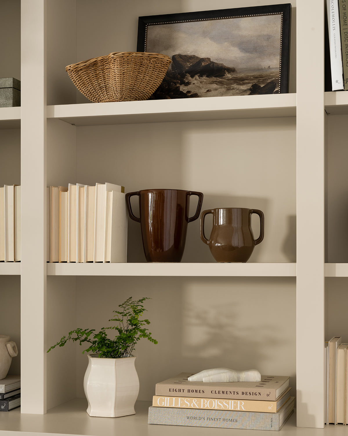 A beige bookshelf displays a framed landscape painting, a woven basket, the Victoire Vase, a white pitcher with greenery, stacked books, and rows of vertically and horizontally arranged books, by Vendixs
