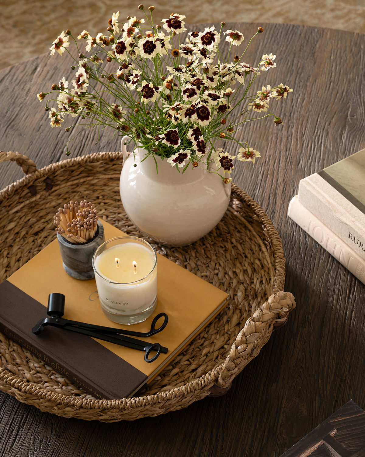 A Salter Woven Tray on a wooden table holds a white vase with daisies, a lit candle, wick trimmer, marble match holder, notebook, and closed book; two stacked books sit next to the tray, by Vendixs