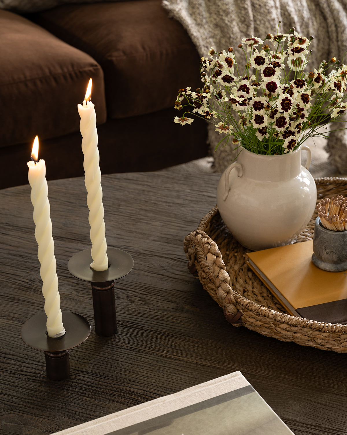 Two twisted white candles in black holders are lit on a dark wooden table. Nearby, a woven tray holds the Rasley Handled Jar with white glaze, a vase of daisies, and stacked notebooks. A brown couch with a textured throw blanket is in the background, by Vendixs