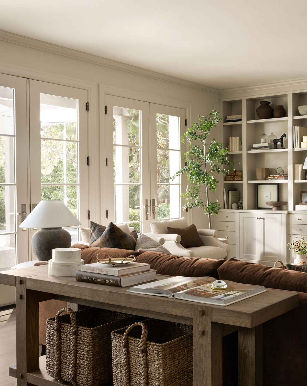 A cozy, sunlit living room with French doors, a brown sofa, and a wooden console table featuring the Salter Woven Square Basket, books, and decor; built-in shelves display vases, books, and plants for a warm, inviting space by Vendixs
