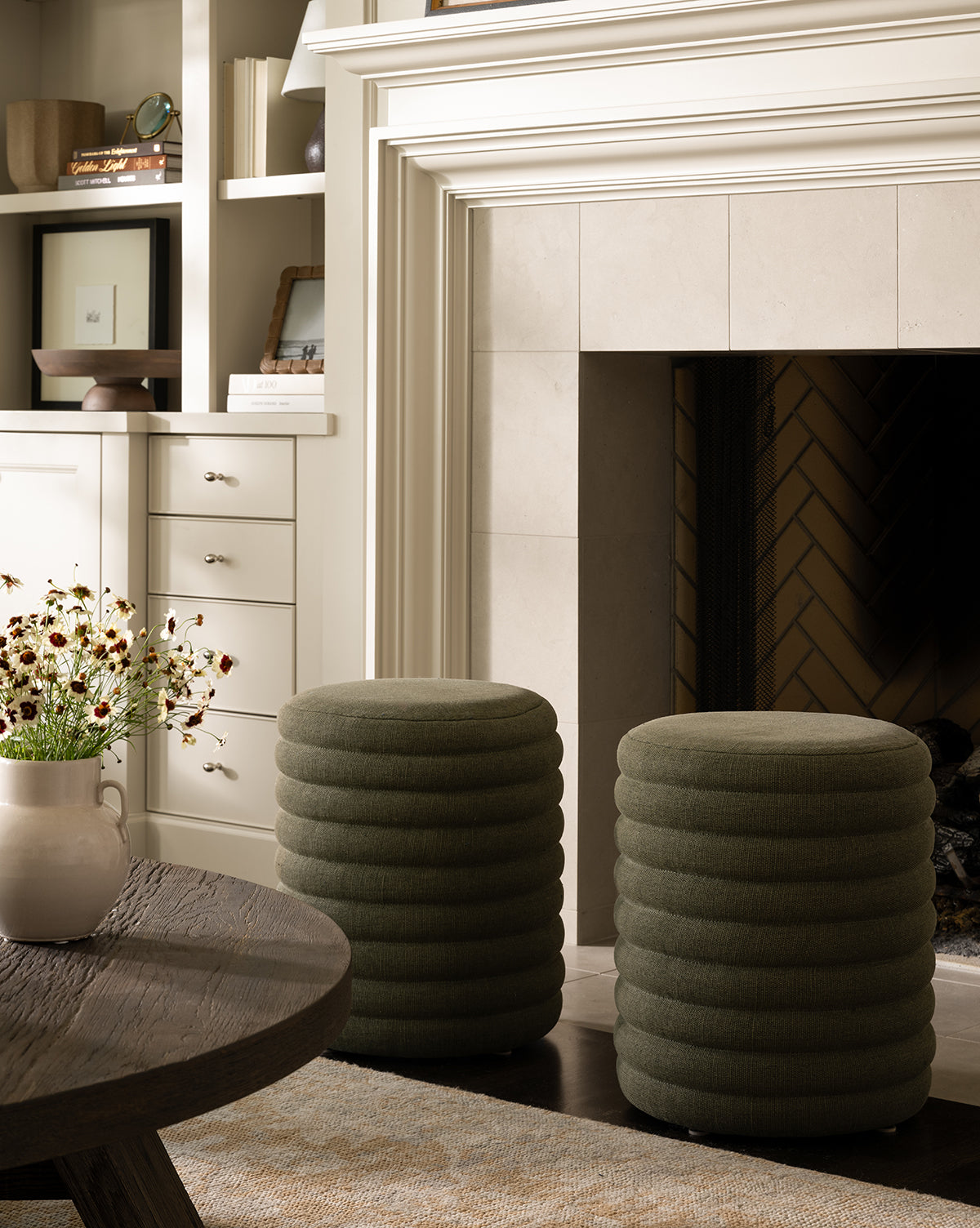 Two round, textured Jefferson Stools by Made Goods in green quilted fabric sit before a white-mantel fireplace. A wooden coffee table with a flower vase and built-in shelves/cabinets can be seen in the background, blending seamlessly with touches from Vendixs