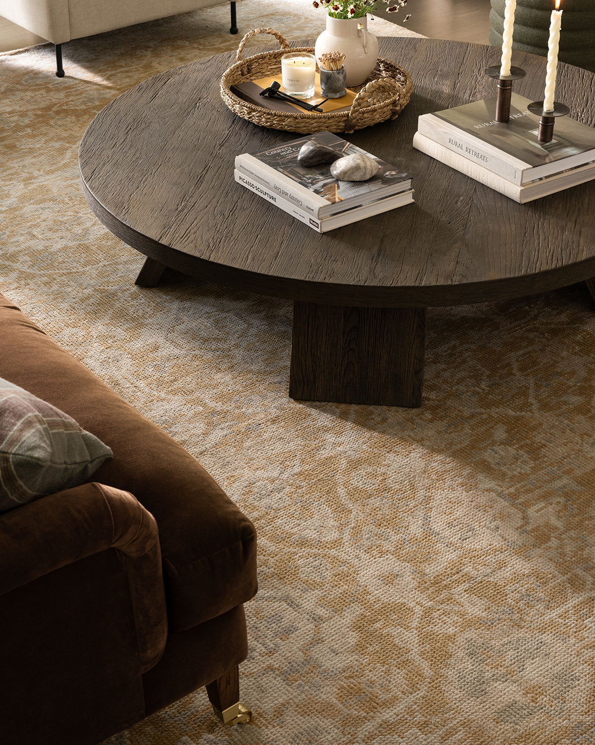 A round, dark wood coffee table topped with books, stones, candles, and a wicker tray sits on the Vanderwood Hand-Knotted Wool Rug near a brown upholstered sofa and cream armchair in a cozy living room, by Vendixs
