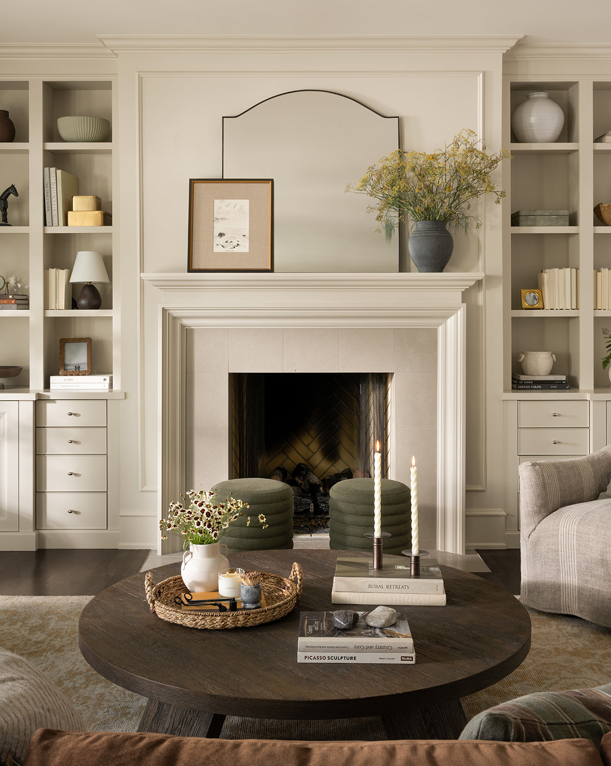 A cozy living room features a fireplace, built-in shelves, and a round dark wood coffee table with books, candles, and a vase. The Willson Wall Mirror with its iron frame enhances the decor as neutral tones and soft lighting create warmth by Vendixs