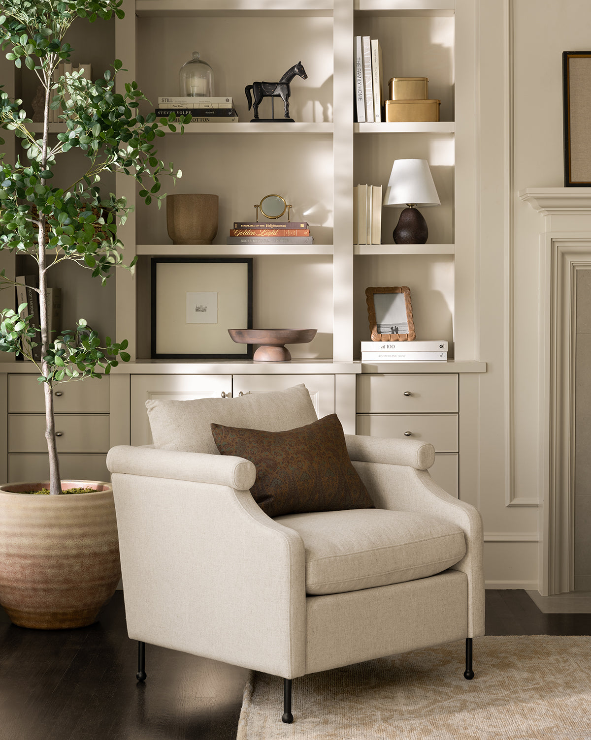 A cozy beige Clegg Lounge Chair by Makerspalm with a brown pillow sits before built-in shelves of books, decor, and a lamp. A large potted plant on the dark wood floor brings vintage charm to this inviting living space. Styled with accents from Vendixs