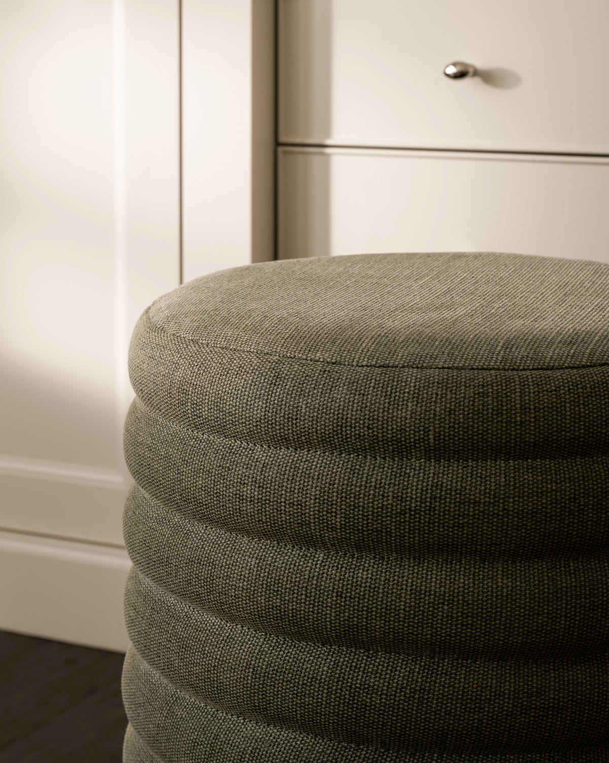 A close-up of the Made Goods Jefferson Stool, featuring a round, textured gray quilted design with horizontal ridges, positioned in front of light-colored cabinets and drawers with metallic knobs. Vendixs