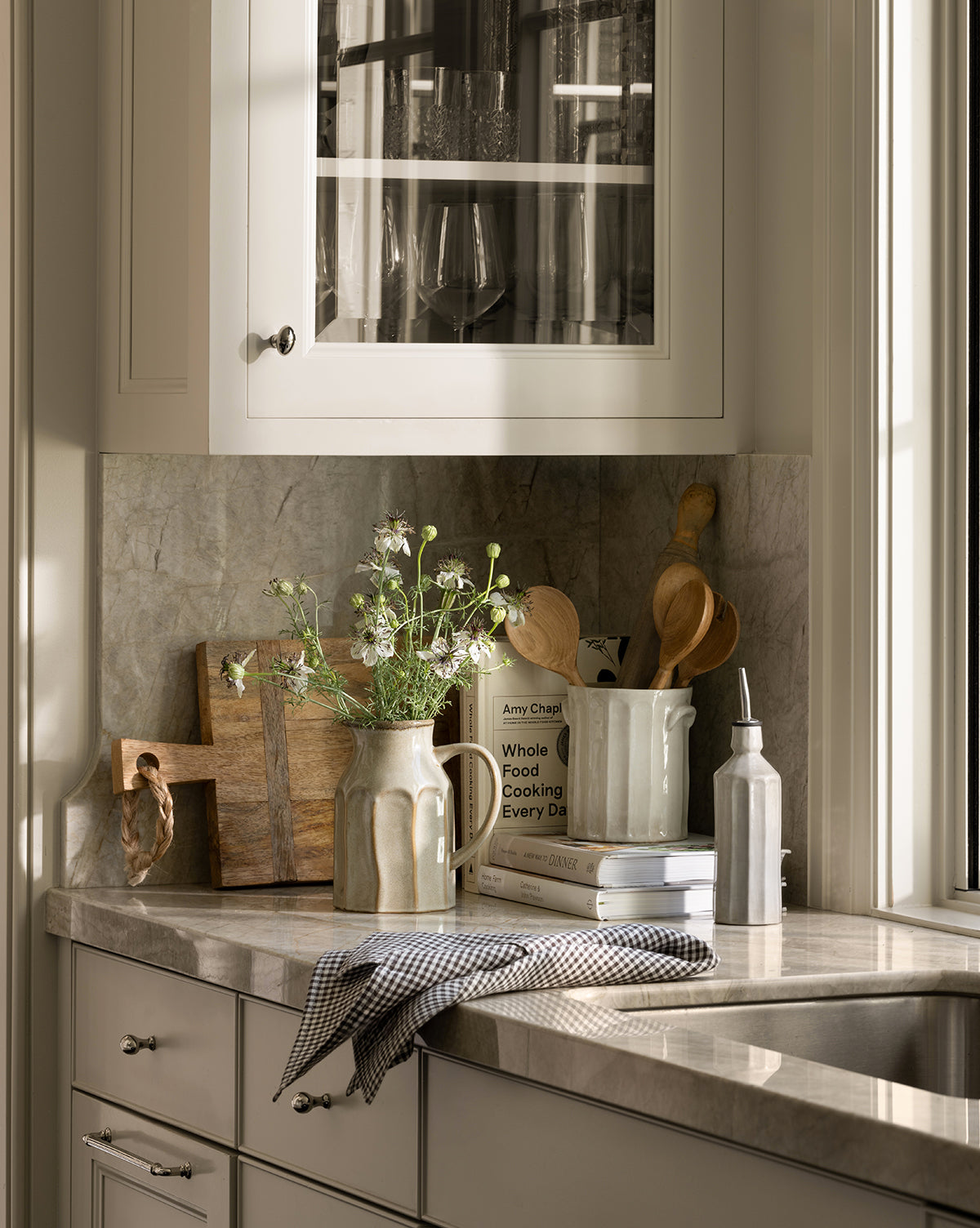 A sunlit kitchen counter features the Westport Linen Tea Towel in brown checks, wildflowers in a vase, cutting boards, wooden utensils in a ceramic jug, cookbooks, a soap dispenser by the sink, and glassware displayed above, by Vendixs