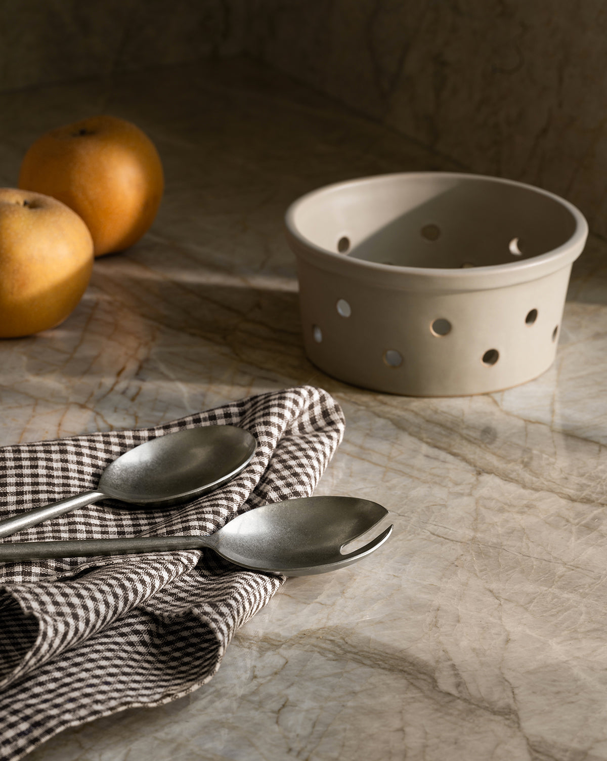 Two metal spoons rest on the Westport Linen Tea Towel atop a marble surface. Warm natural light casts soft shadows, highlighting the towel alongside two apples and a beige ceramic basket with holes, by Vendixs