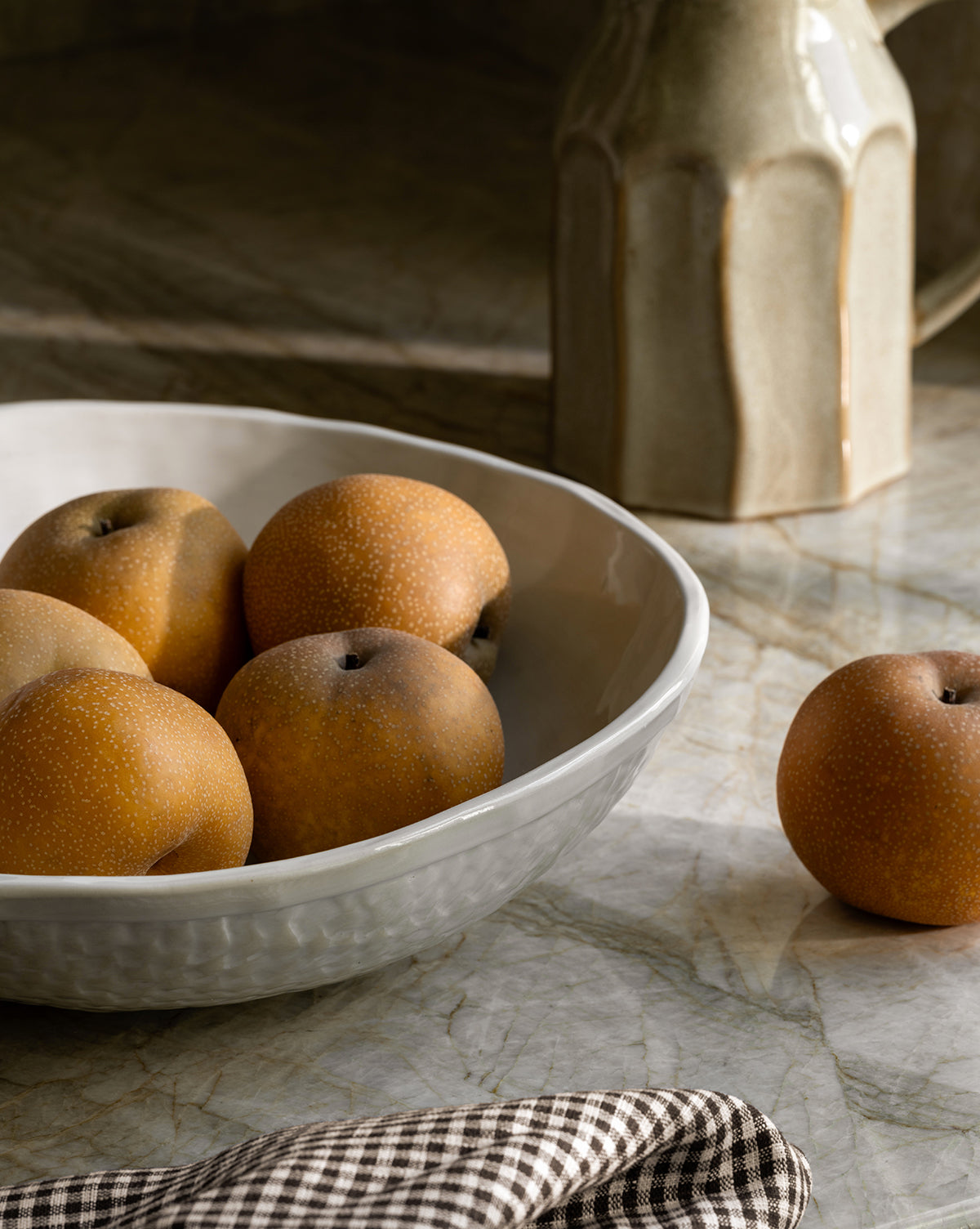 The Dion Serving Bowl, finished in white ceramic glaze, holds five Asian pears on a marble surface with another pear nearby. A beige jug and checked cloth add elegance to the tranquil arrangement by Vendixs