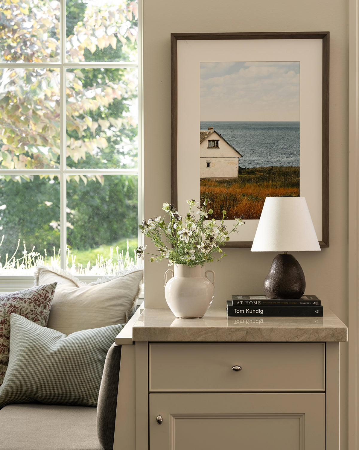 A cozy corner features a cushioned bench by the window, a vase of white flowers, books on a beige cabinet, and the Vedruna Table Lamp with its brown ceramic base and tapered white shade beneath a coastal house photo by Vendixs