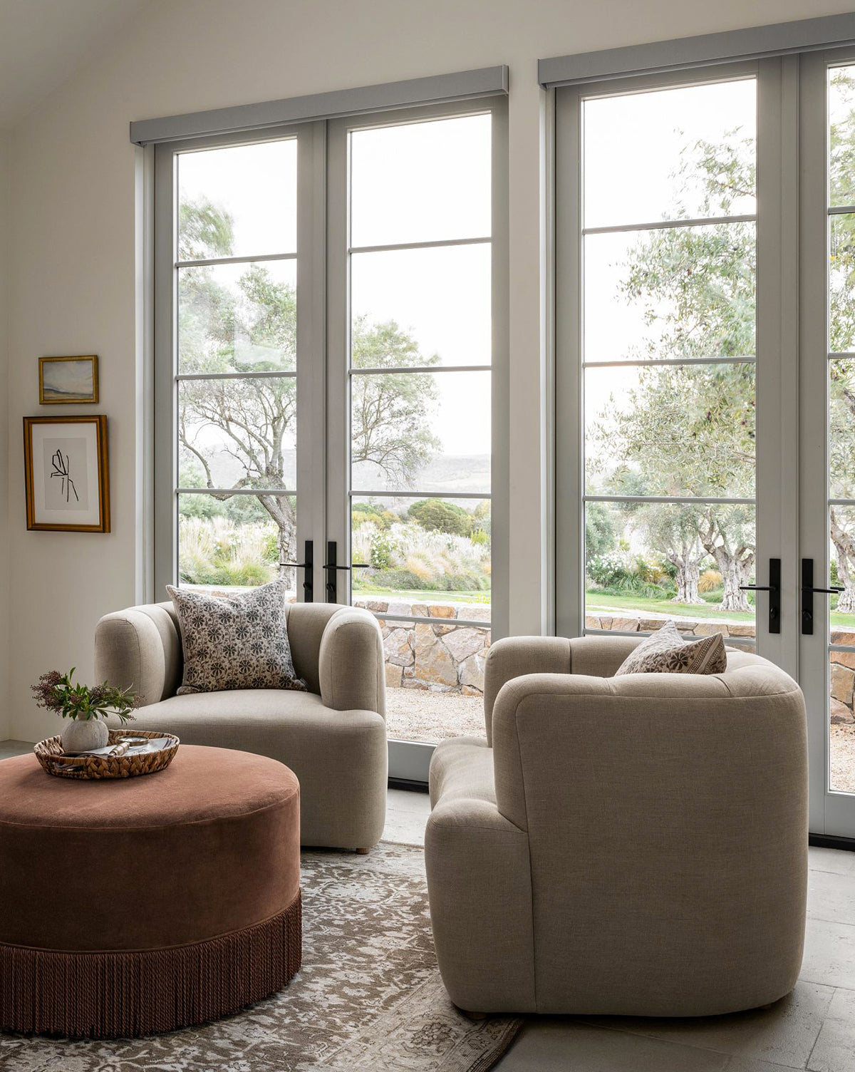 Two beige Community Ines Lounge Chairs with patterned cushions face a round brown ottoman in a bright room with large windows, modern decor, a side table, framed wall art, and views of a lush garden. Vendixs