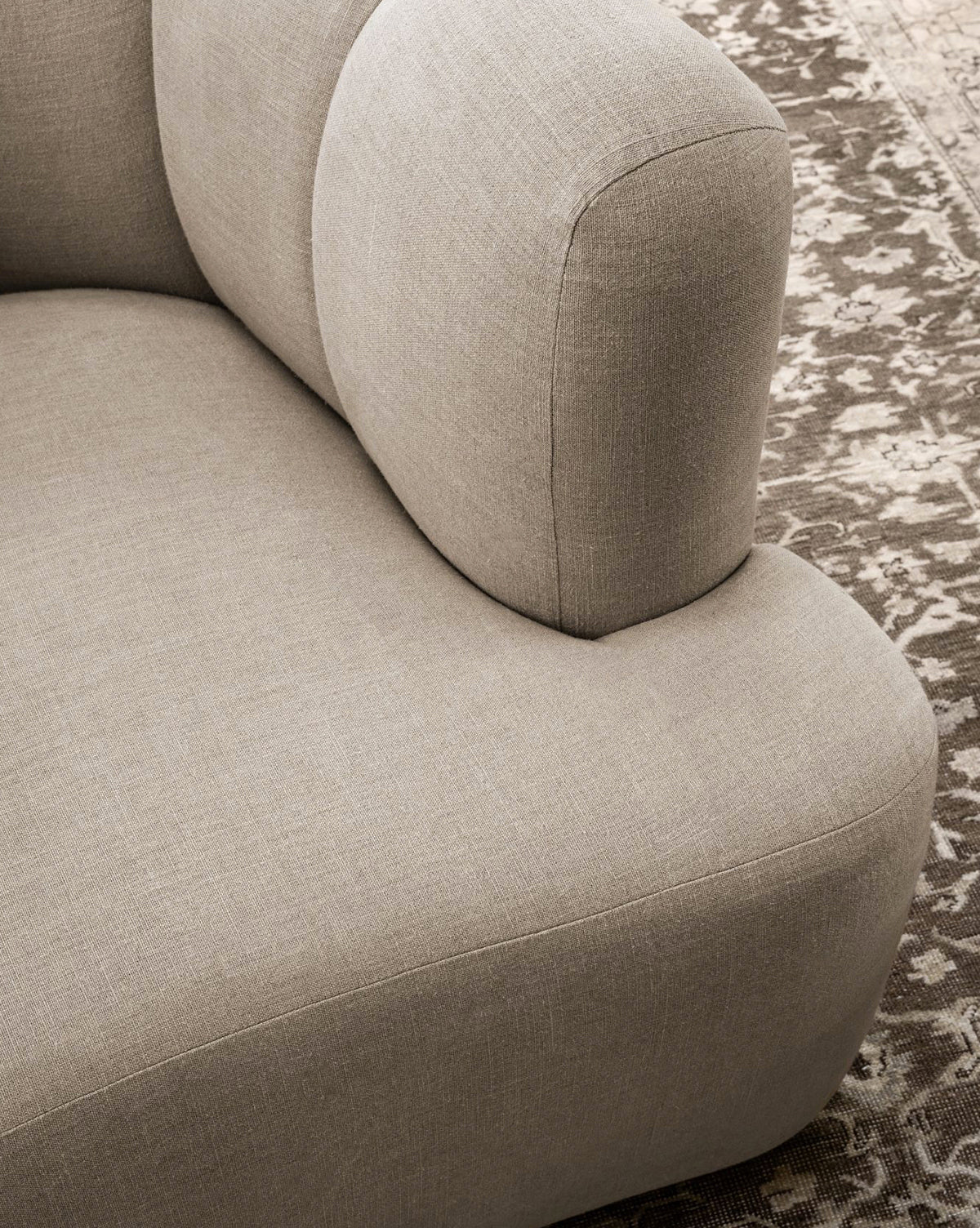 Close-up of a light beige upholstered sofa with rounded, cushioned armrest and backrest, placed on a brown patterned rug beside the modern Ines Lounge Chair by McGee & Co.