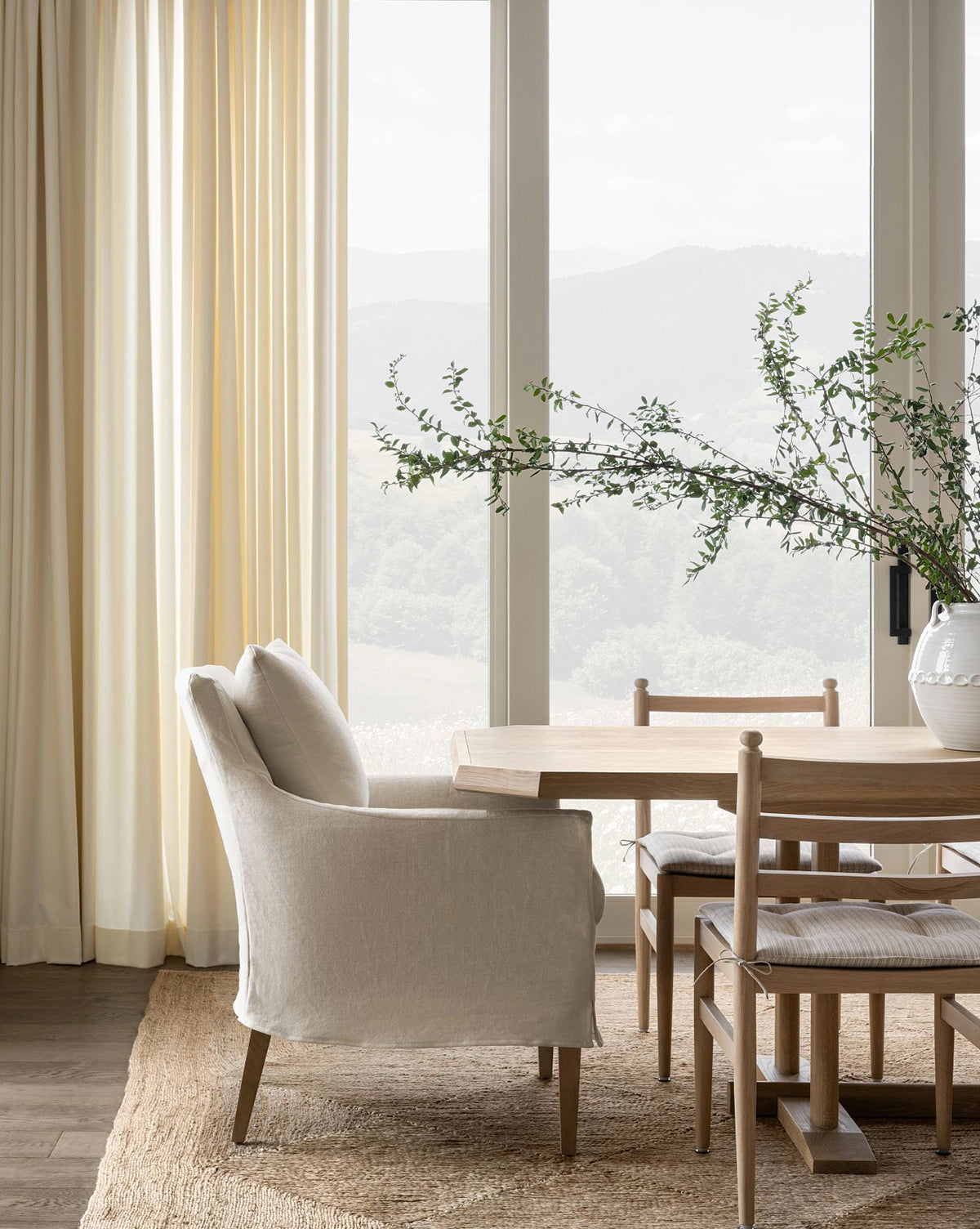 A bright dining area features a light wood table, the Sascha Slipcover Dining Chair, woven chairs, an upholstered armchair, a large vase with green branches, neutral curtains, a jute rug, and big windows with misty mountain views by McGee & Co.