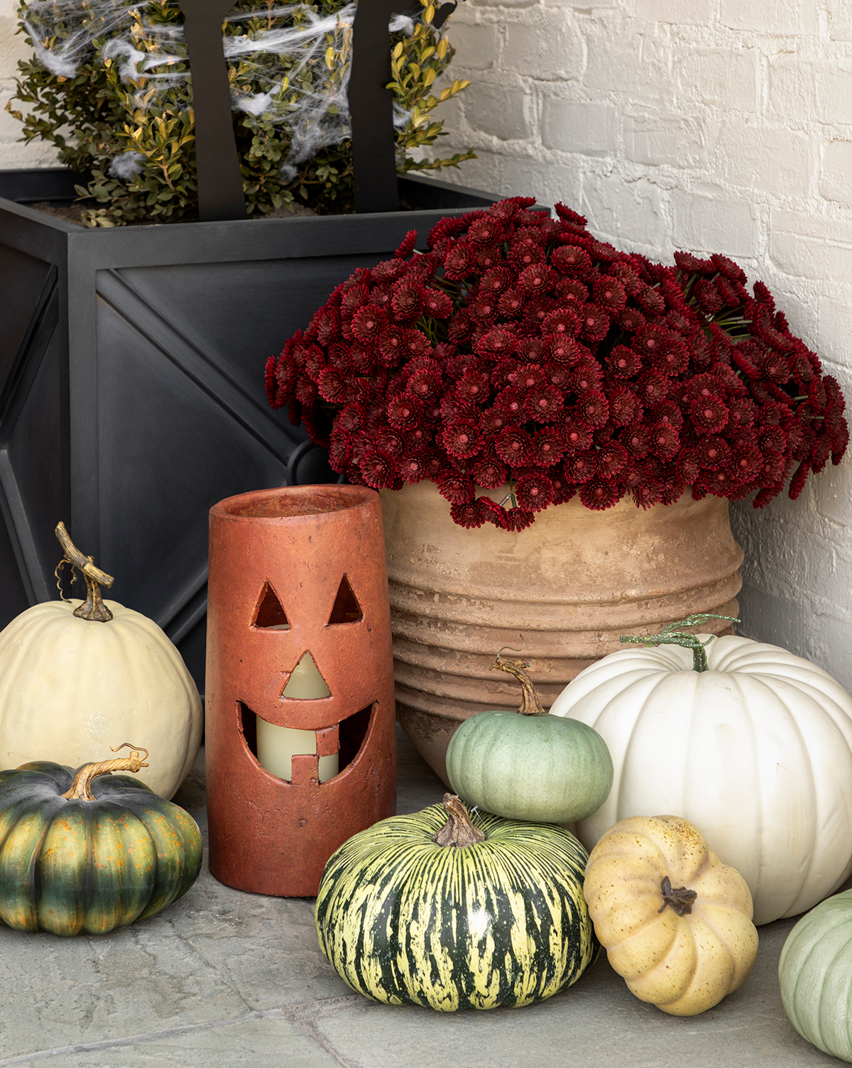A fall porch display featuring white and green pumpkins, a terracotta jack-o-lantern candle holder, a black planter with greenery, and a pot of Faux Mum Drop-In blooms set against a light brick wall for inviting seasonal decor, by Vendixs