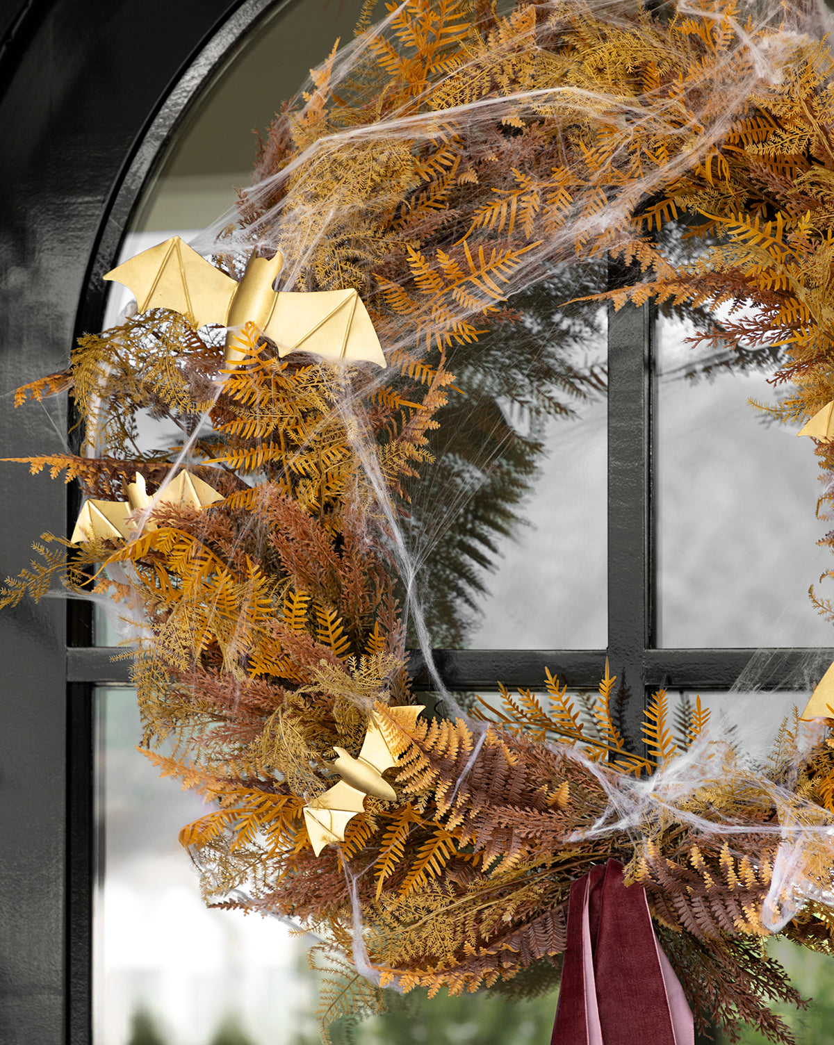 The 30 Faux Golden Fern Wreath hangs on a black door, featuring dried autumn leaves, gold origami bats, artificial cobwebs, and a burgundy ribbon. Ideal for fall entryway decor, by Vendixs