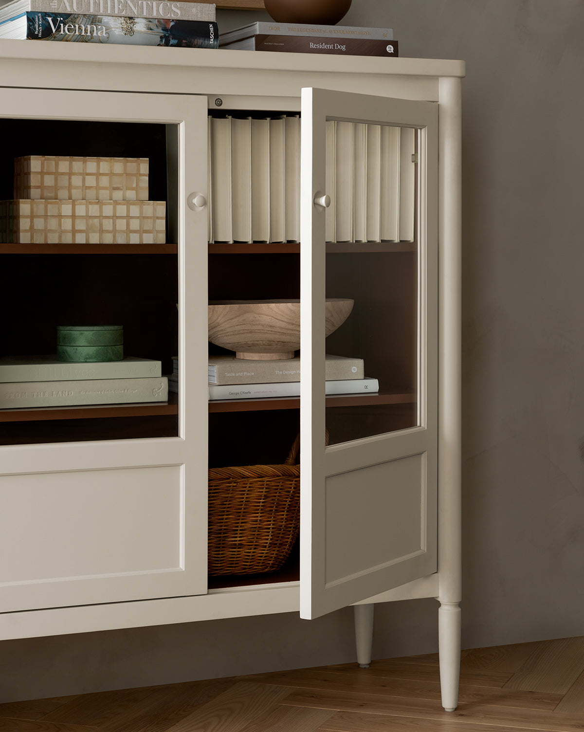 A bone white Highgate Cabinet with glass doors stands partly open, displaying shelves with books, a wooden bowl, a woven basket, and decorative boxes. Several books are stacked on top of this elegant Highgate Cabinet by Vendixs