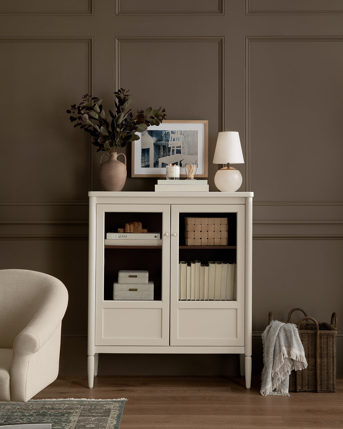 A cream cabinet with books and boxes stands against a brown paneled wall. On top are the Terri Round Table Lamp with an alabaster base, framed art, stacked books, and a vase of leafy branches. A beige armchair and basket with blanket are nearby, by Vendixs