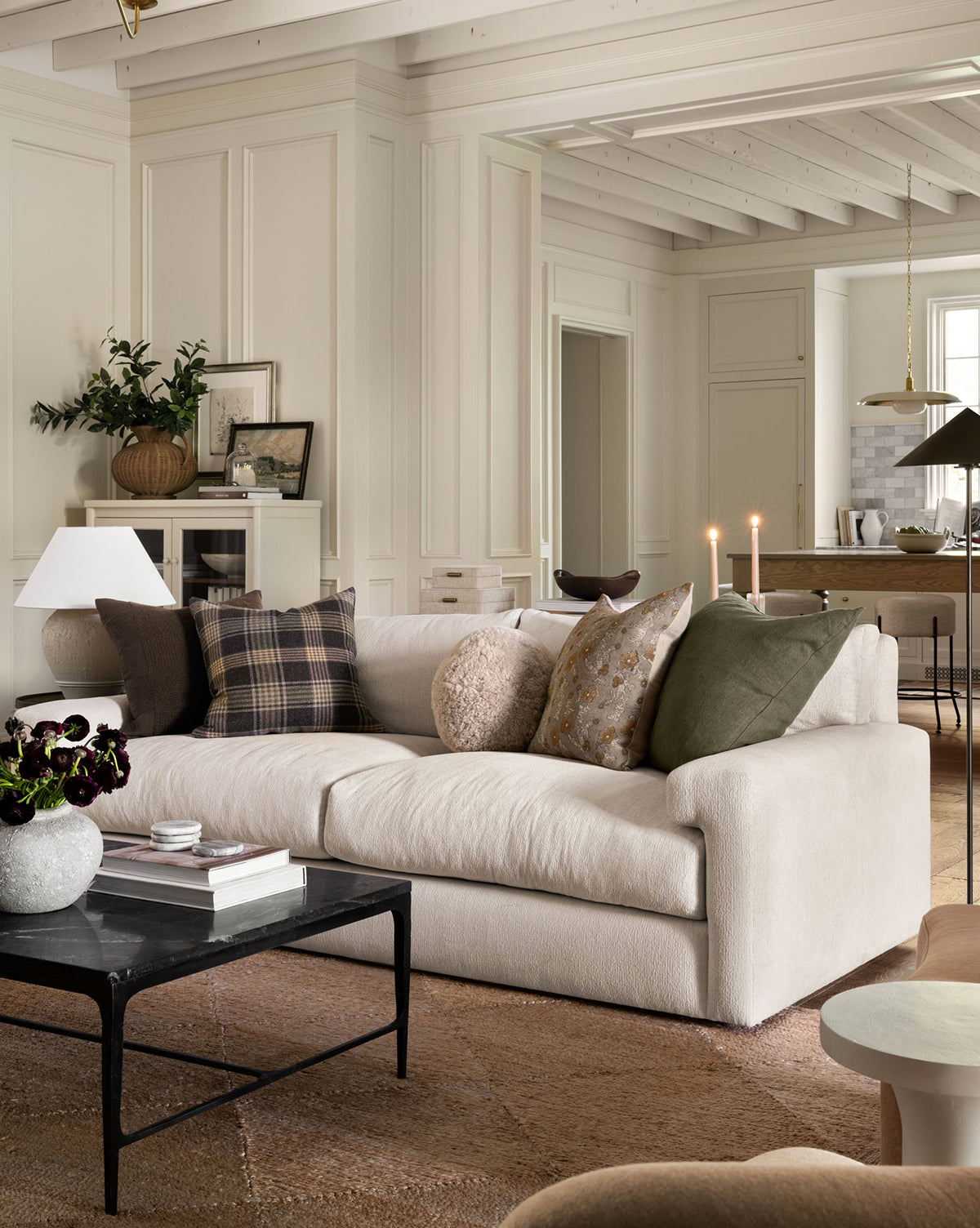 A cozy, modern living room showcases the Community Beckstead Sofa 92 with assorted throw pillows, a black coffee table, and candles. Neutral tones and layered textures create a warm, inviting vibe. The dining area appears in the background, featuring accents from Vendixs