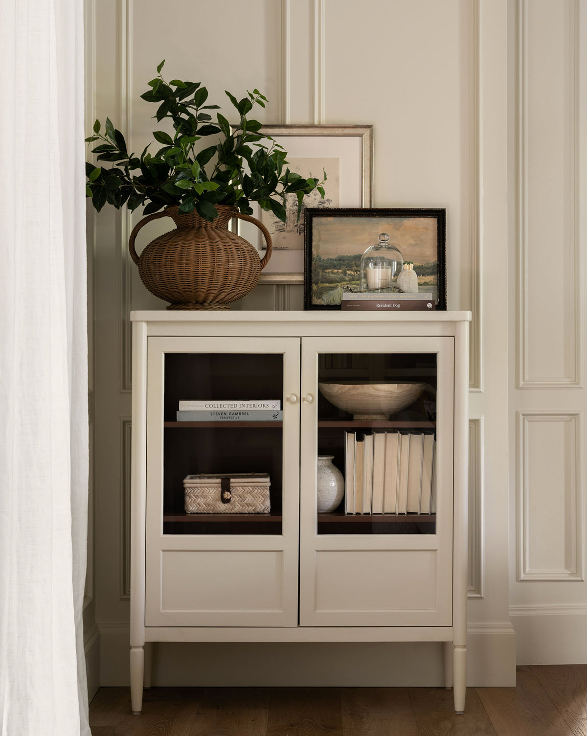 A bone white Highgate Cabinet with glass doors showcases books, a woven box, and decor. On top are framed art, a wicker vase with green branches, and more books. The scene is bright and elegant with Victorian-inspired charm, by Vendixs