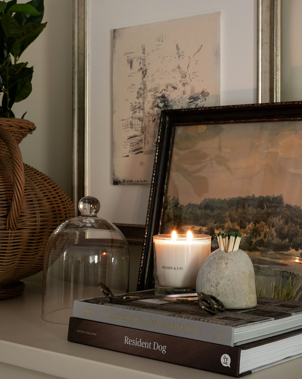A cozy scene with a lit Palo Santo & Oakmoss Candle, stone match holder, art books, framed artwork, a wicker vase of greenery, and a glass cloche on the shelf—warm, inviting, and tastefully arranged by Vendixs