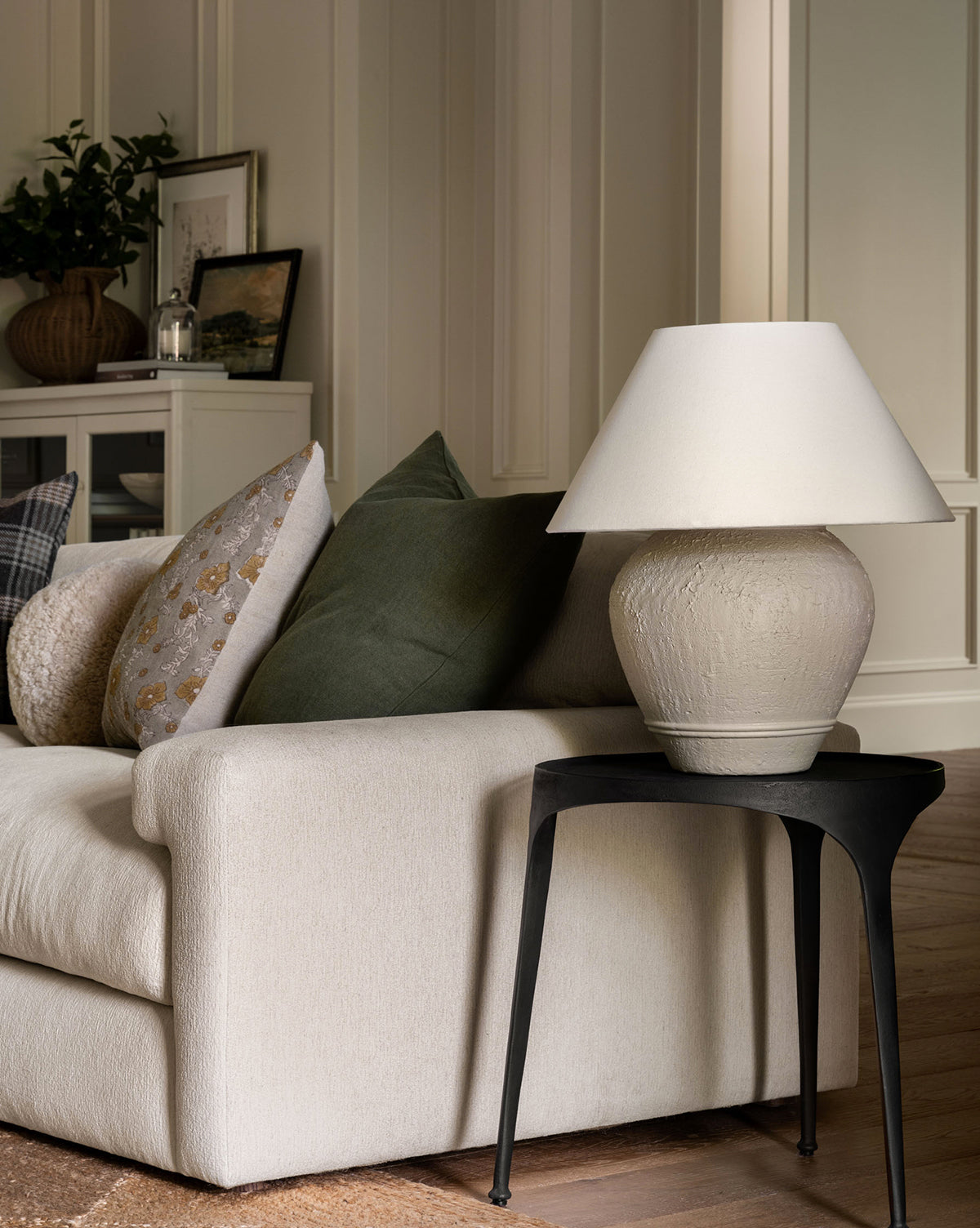 A cozy living room corner with a beige sofa and assorted pillows sits next to a black side table topped with the Abbott Ceramic Table Lamp, featuring a round ceramic base and white linen shade, creating a warm, softly lit atmosphere by Vendixs