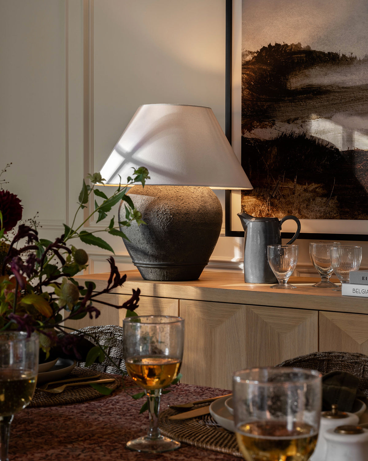 A cozy dining room features kitchen touches—a Levine Ceramic Pitcher in dark gray glaze and glassware among flowers on the table, with a textured lamp and framed art accenting a wooden sideboard under warm ambient lighting, by Vendixs