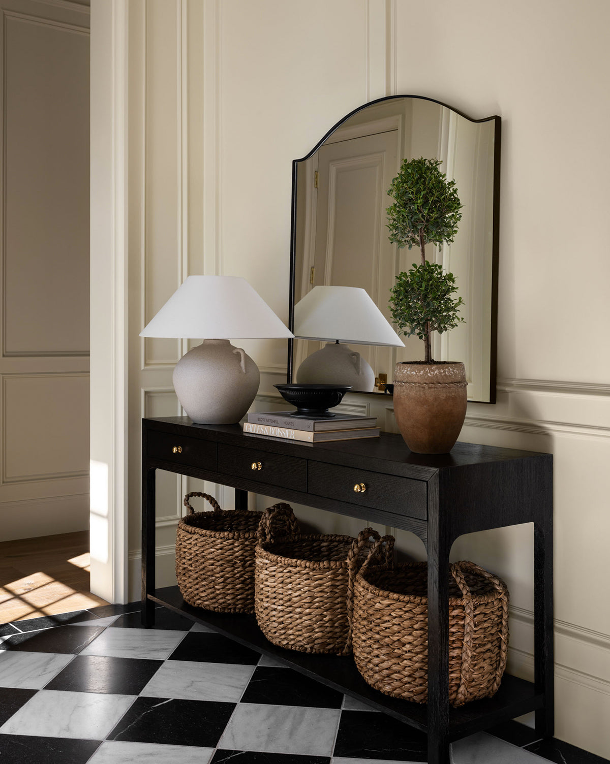 A black console table with drawers displays a white lamp, a bowl, and a potted topiary, with three woven baskets below. The Willson Wall Mirror’s iron frame reflects against cream-paneled walls and checkered black-and-white flooring by Vendixs