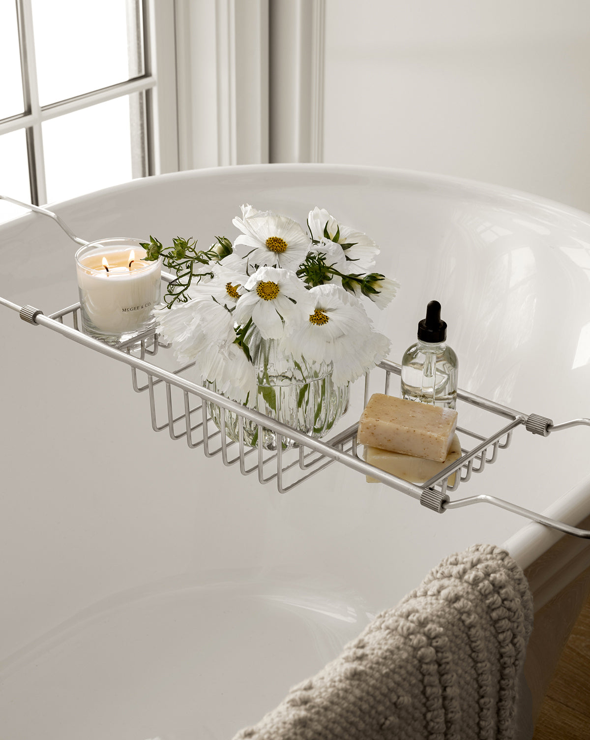 A bathtub with a metal tray holds a lit Salt & Sea Candle, a glass jar of white flowers, a bar of soap, a dropper bottle, and a textured gray towel draped over the edge near a sunlit window, by Vendixs