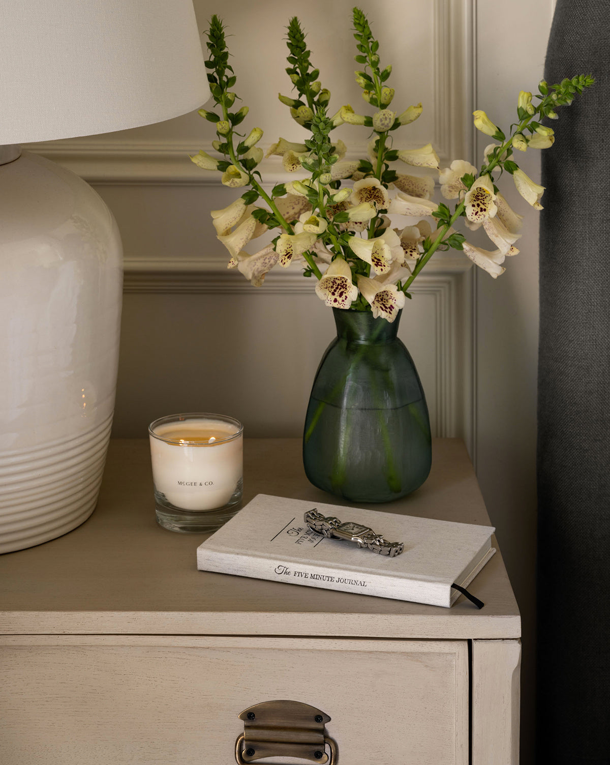 A small beige nightstand holds a green vase of pale yellow flowers, a lit Citrus & Birch Candle, and a closed journal with a silver pen. Nearby are part of a white lamp and a dark curtain, by Vendixs