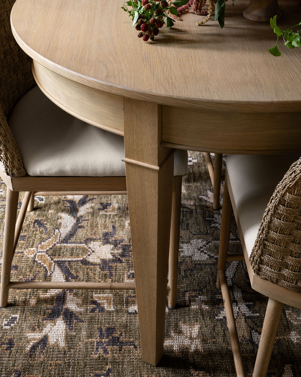 A round wooden dining table with woven chairs and beige cushions sits atop the Wayland Hand-Knotted Wool Rug. A small plant brings a lively accent to this cozy, inviting setting, by Vendixs