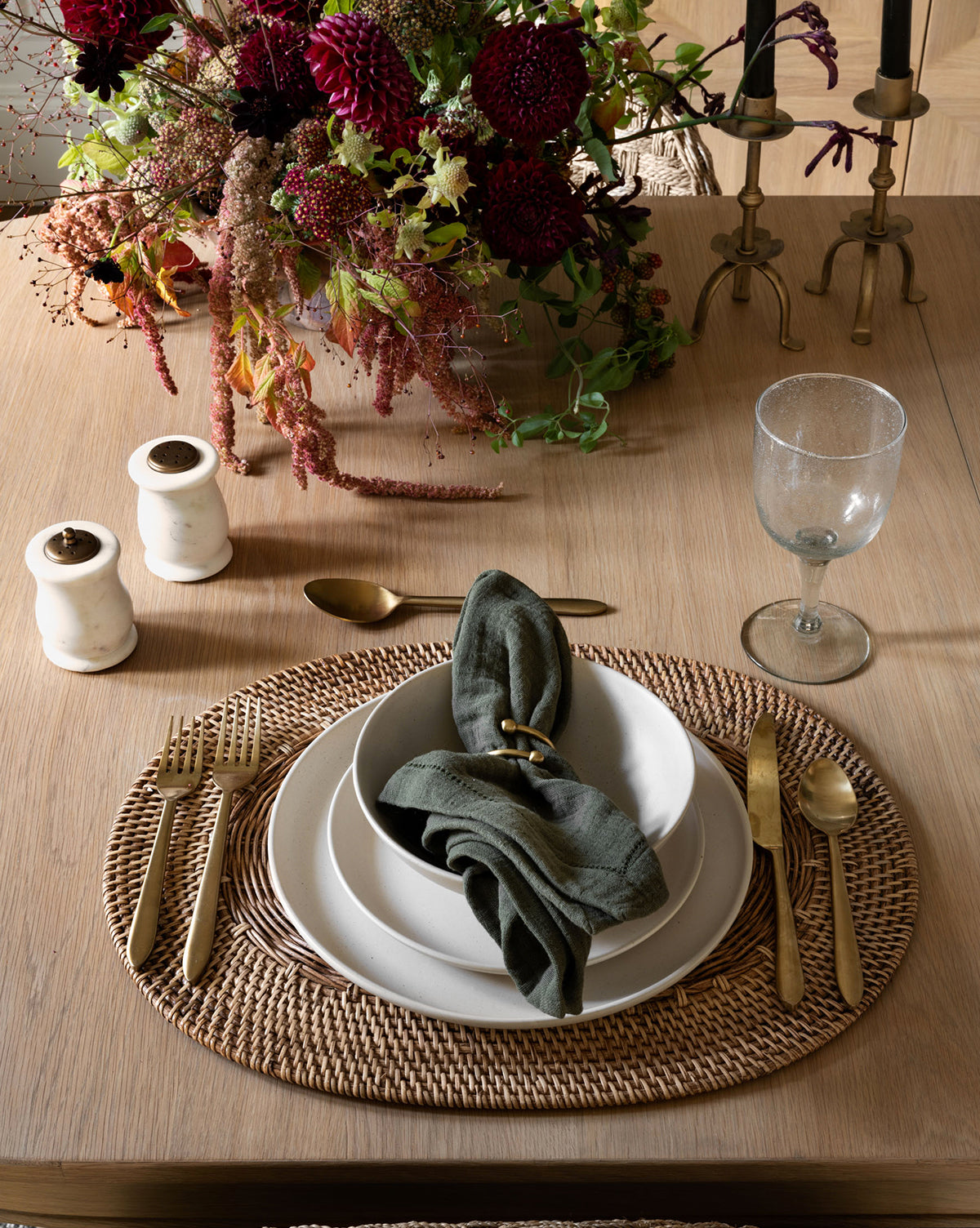 A table setting with gold flatware, white plates, a green napkin, a clear goblet on the Davin Placemat. Salt and pepper shakers, brass candlesticks, and a flower arrangement complete the elegant look, by Vendixs