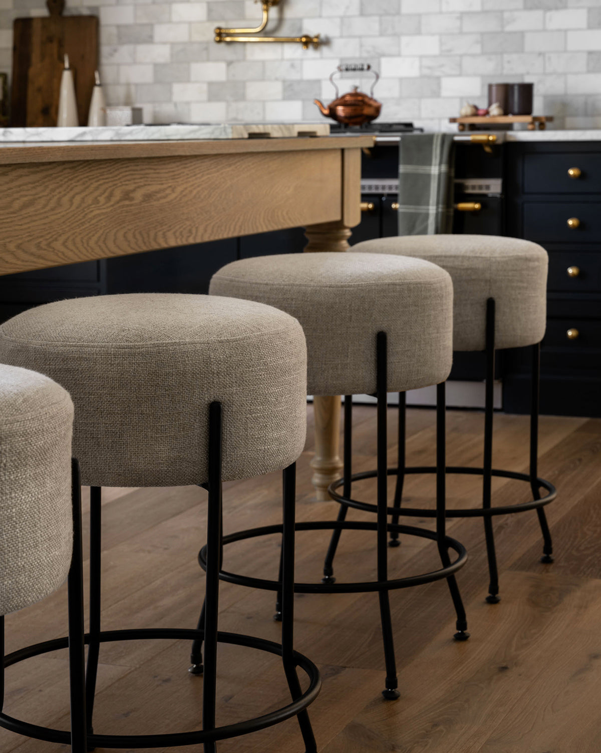 Three Makerspalm Evander Counter Stools with black metal legs are lined up at a wooden kitchen island, complementing a modern space with tile backsplash, dark cabinetry featuring brass hardware, and wooden flooring, by Vendixs