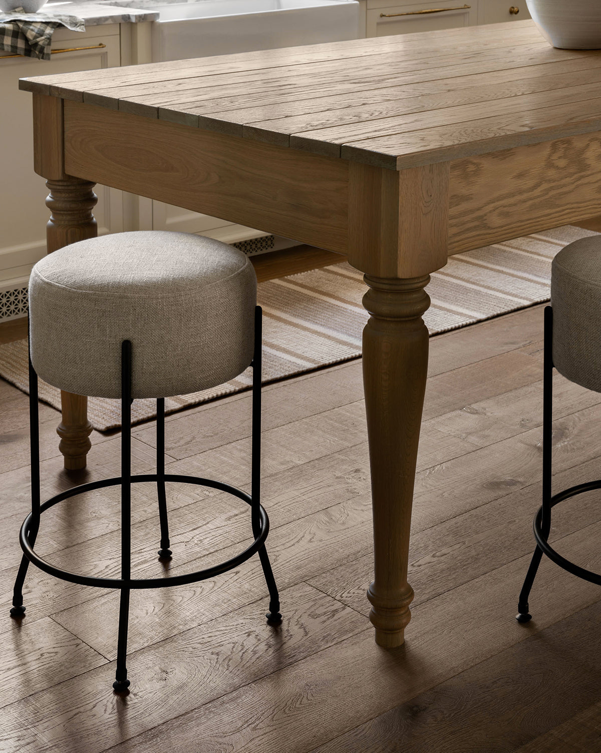 A close-up of a wooden kitchen table with turned legs, two Makerspalm Evander Counter Stools with metal legs, and a striped rug on hardwood in a bright room—ideal for modern kitchen seating. Vendixs