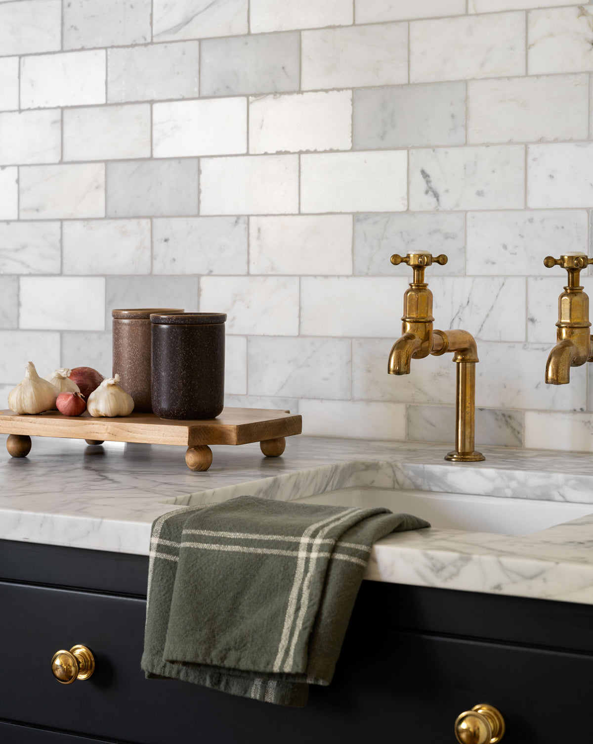 A marble kitchen countertop features a gold faucet, green striped towel, and the Cillian Pedestal Board in pine wood displaying jars and garlic cloves. White marble tile backsplash and gold drawer knobs complete the look by Vendixs