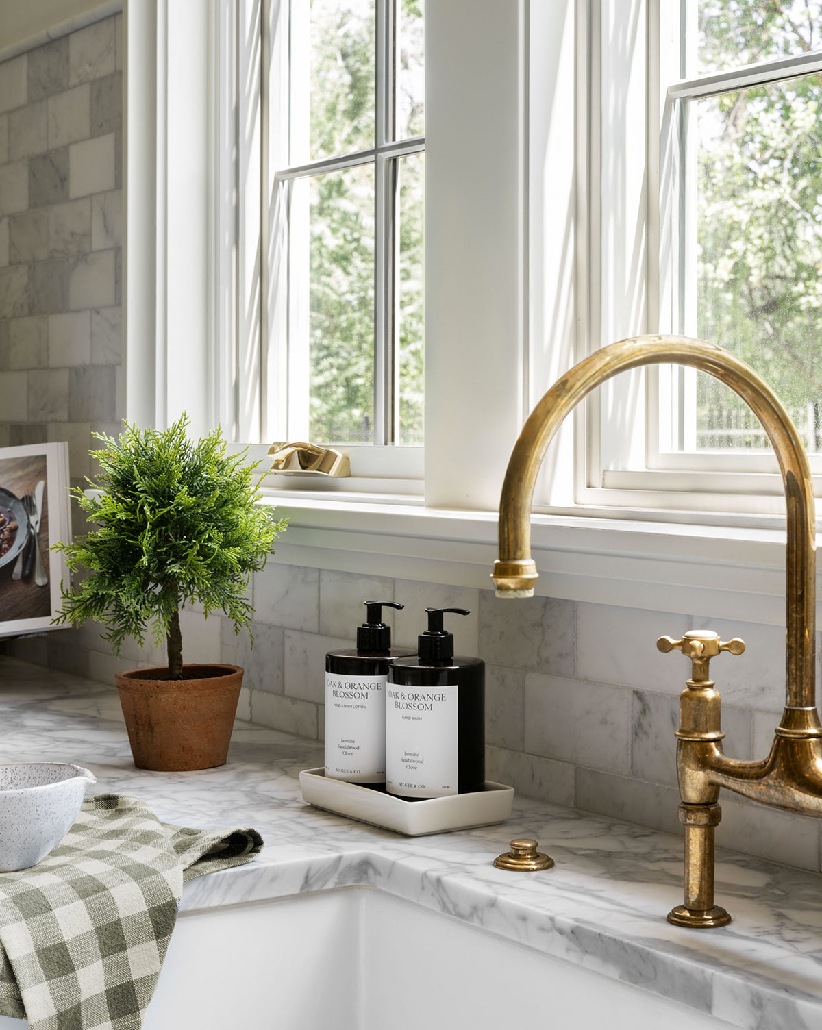 Close-up of a kitchen sink area with a brass faucet, soap bottles on a tray, and a checkered towel on marble. A 15.5" Cypress Faux Potted Topiary in a terracotta pot adds greenery beside large windows, by Vendixs
