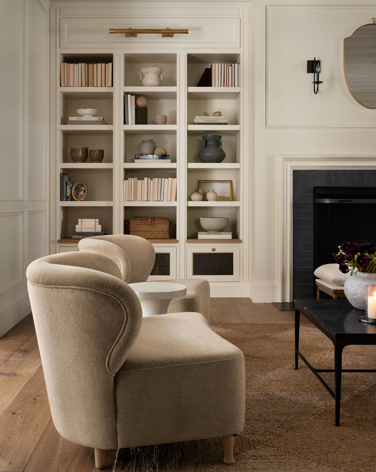Two Carmella Slipper Chairs (Ready to Ship) in beige face a black coffee table in a cozy living room featuring a built-in bookshelf, dark fireplace, neutral rugs, and soft lighting for a warm, inviting atmosphere, by McGee & Co.