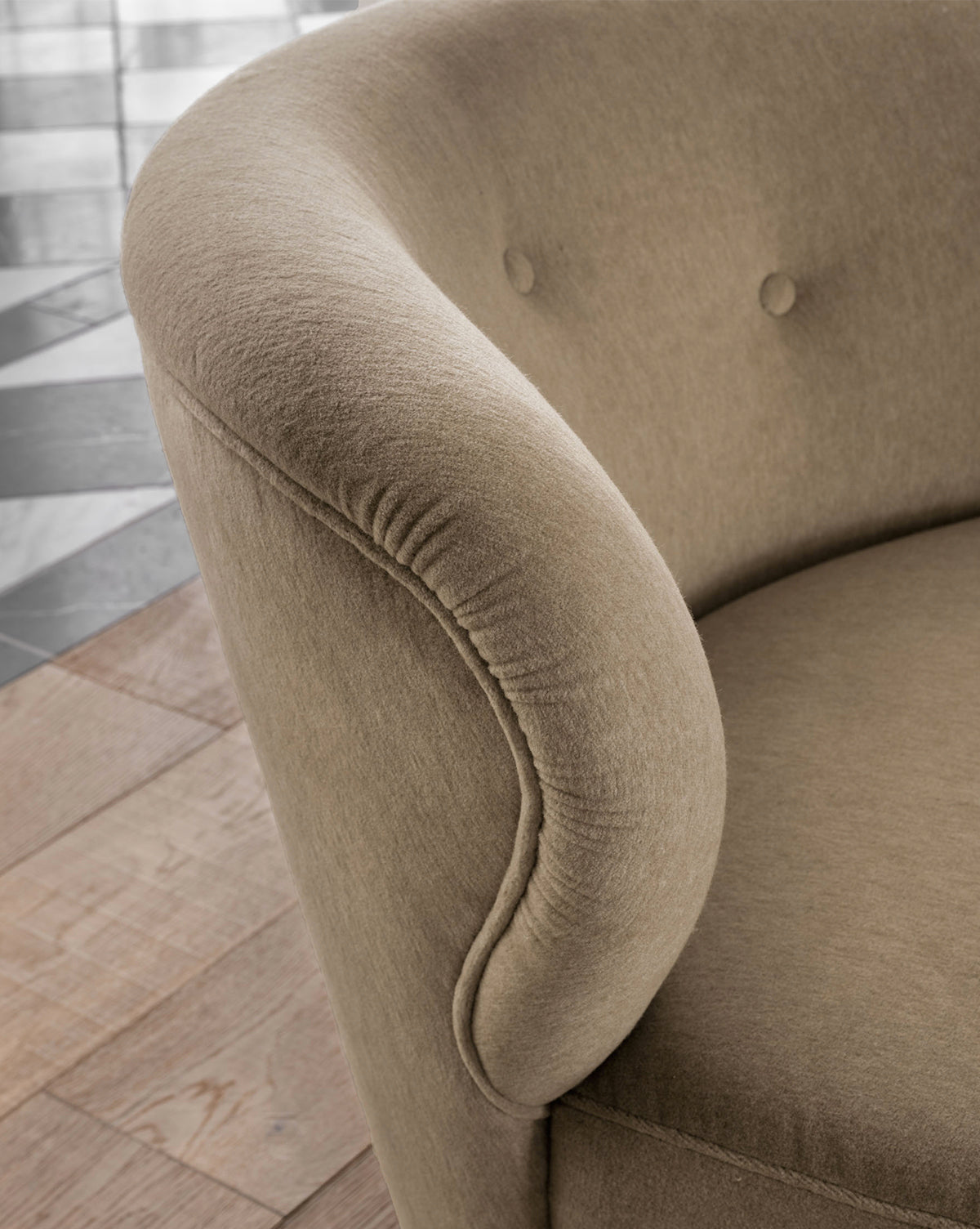 Close-up of the Carmella Slipper Chair (Ready to Ship) featuring a light brown hue, rounded arms, and button-tufted camel upholstery backrest, shown on a wooden floor with black and white tiles in the background by Vendixs