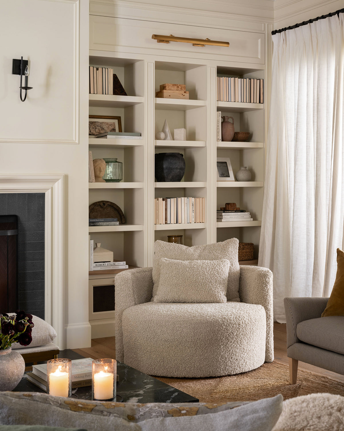 A cozy living room corner features the Banbury Swivel Chair by Rowe Fine Furniture, Inc. in front of built-in bookshelves. Two lit candles rest on a dark coffee table, with soft light filtering through sheer curtains—McGee & Co.