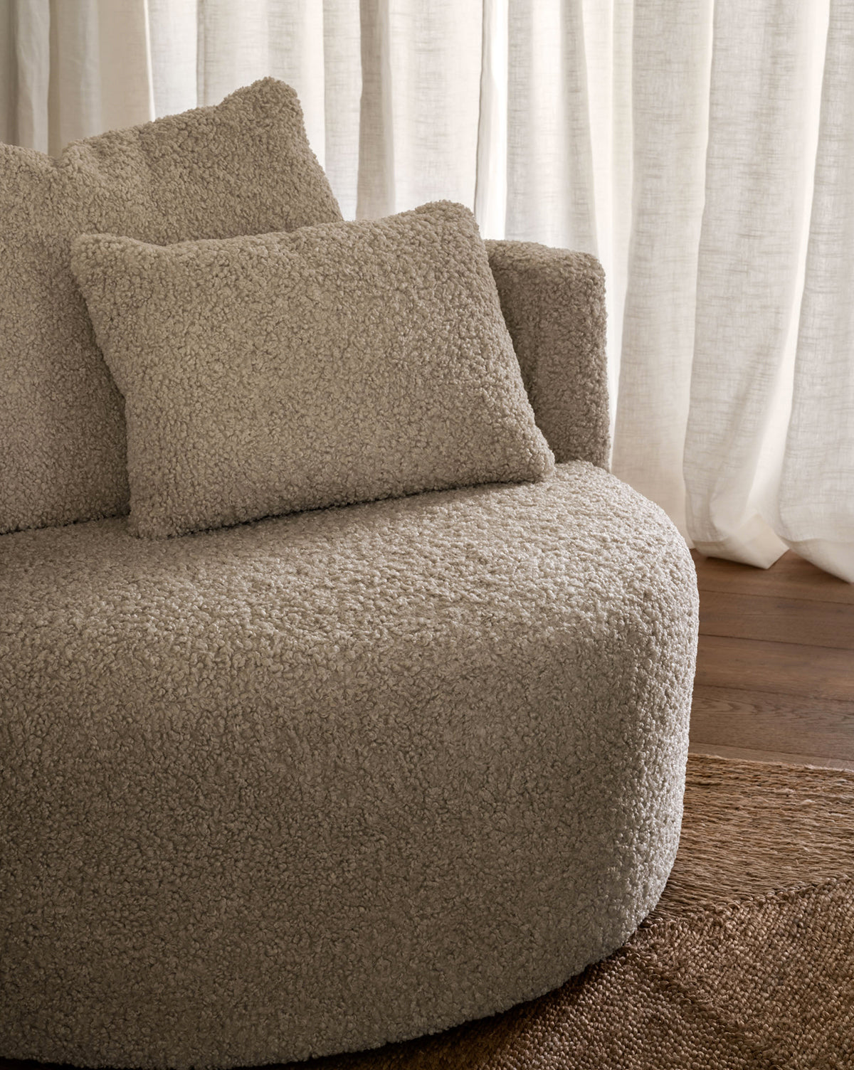 A close-up of the Banbury Swivel Chair, featuring a round, beige boucle design with two matching textured cushions, placed on a woven rug with sheer curtains behind, by McGee & Co.
