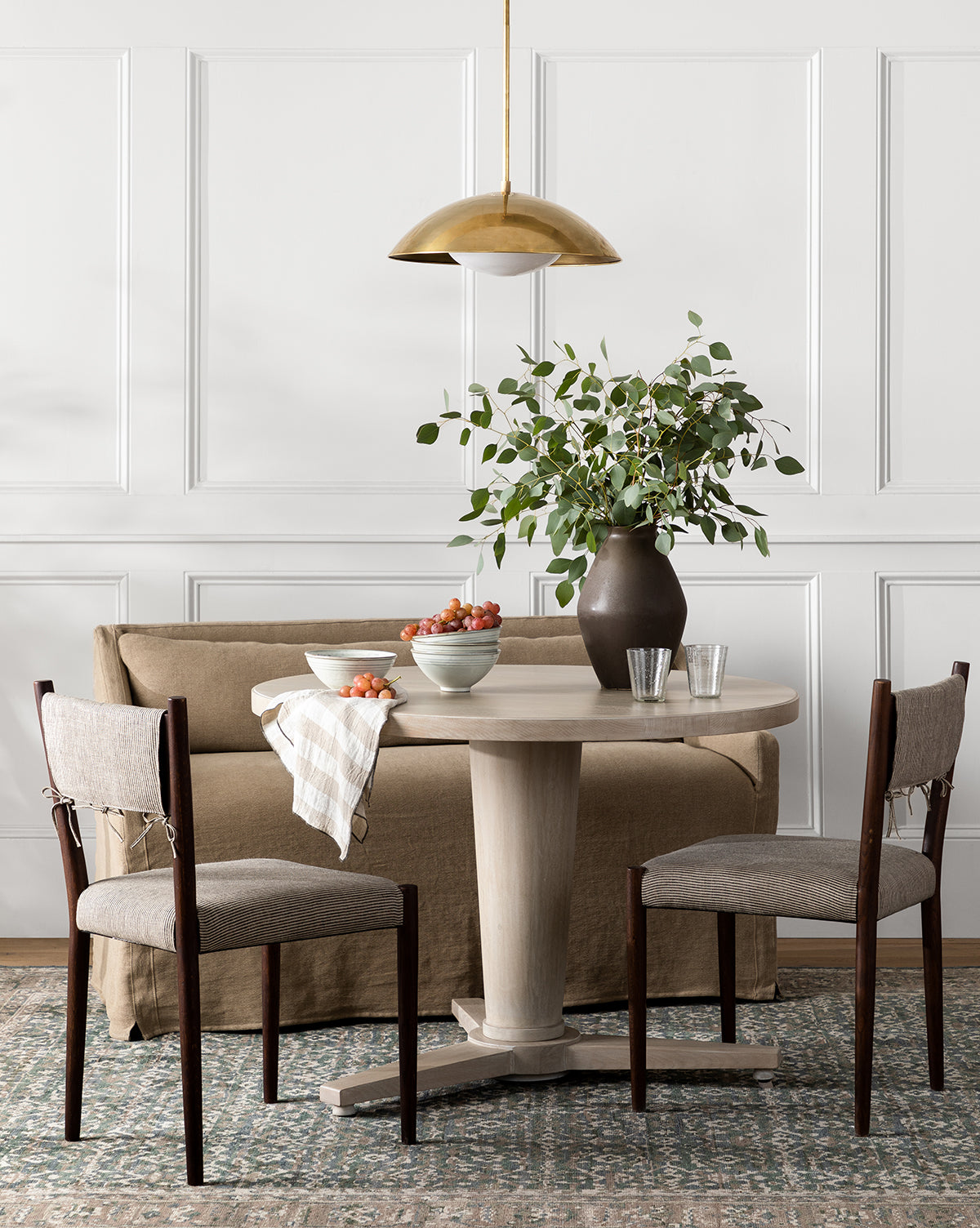 A round wooden dining table with two chairs, a beige sofa, and the Olivier Slipcover Dining Bench is set with bowls of strawberries, glasses, and a dark vase with green branches. A gold pendant light hangs above on a patterned rug by a paneled wall, by McGee & Co.
