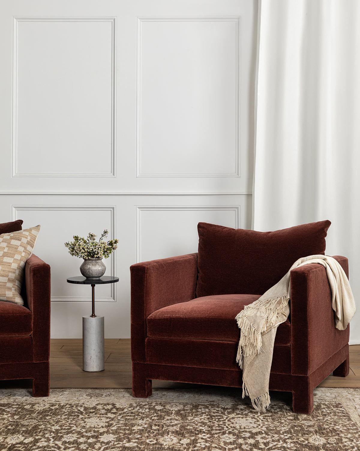 A cozy room features the Makerspalm Appoline Lounge Chair, upholstered in plush dark red with a beige throw. A small side table with a flower vase, neutral walls, white curtains, and a patterned rug complete the scene—McGee & Co.