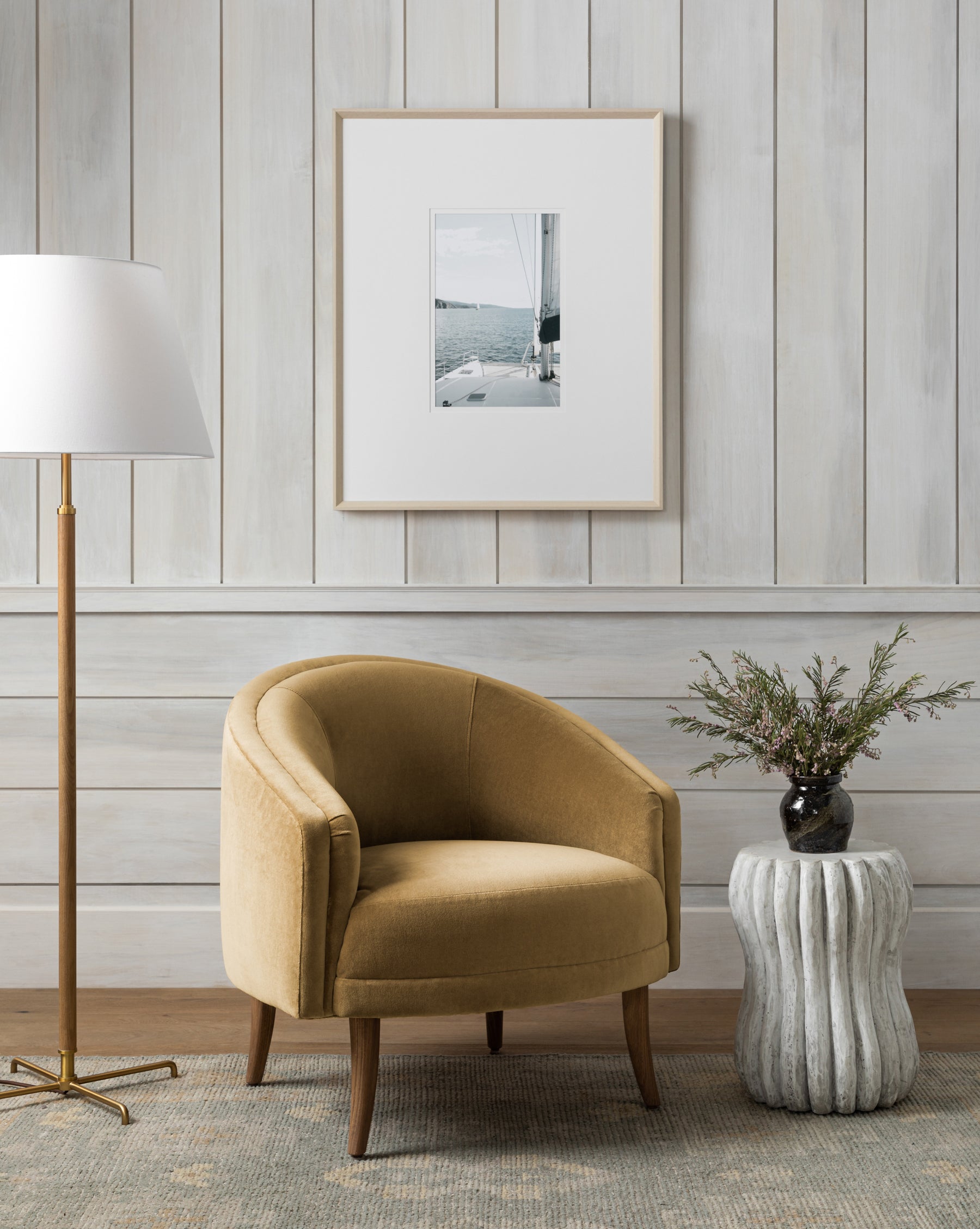 A cozy corner featuring the Marguerite accent chair in gold velvet, paired with a tall white floor lamp, a ribbed round side table topped with greenery in a dark vase, and a sailboat photo on white wood-paneled walls by McGee & Co.