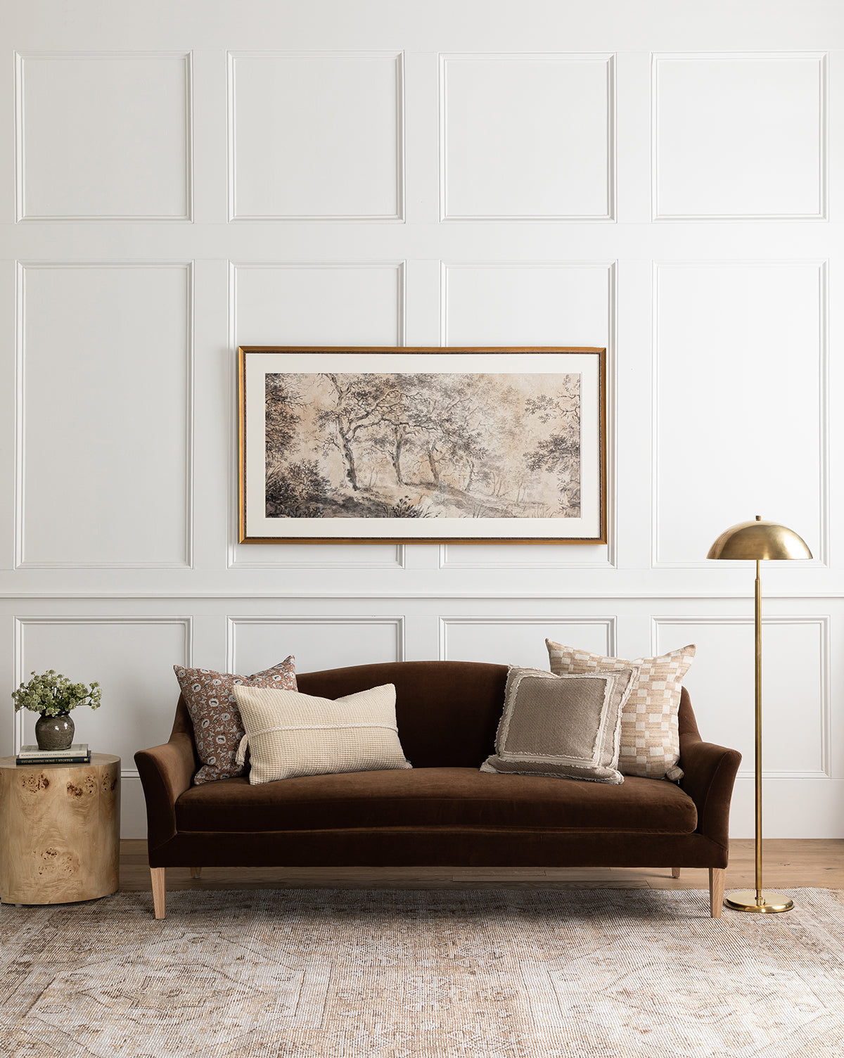 A cozy living room with a dark brown velvet sofa, patterned and neutral pillows, a side table with flowers, brass floor lamp, framed landscape art on a white paneled wall, and the Charlot Hand-Knotted Rug adding character underfoot, by Vendixs