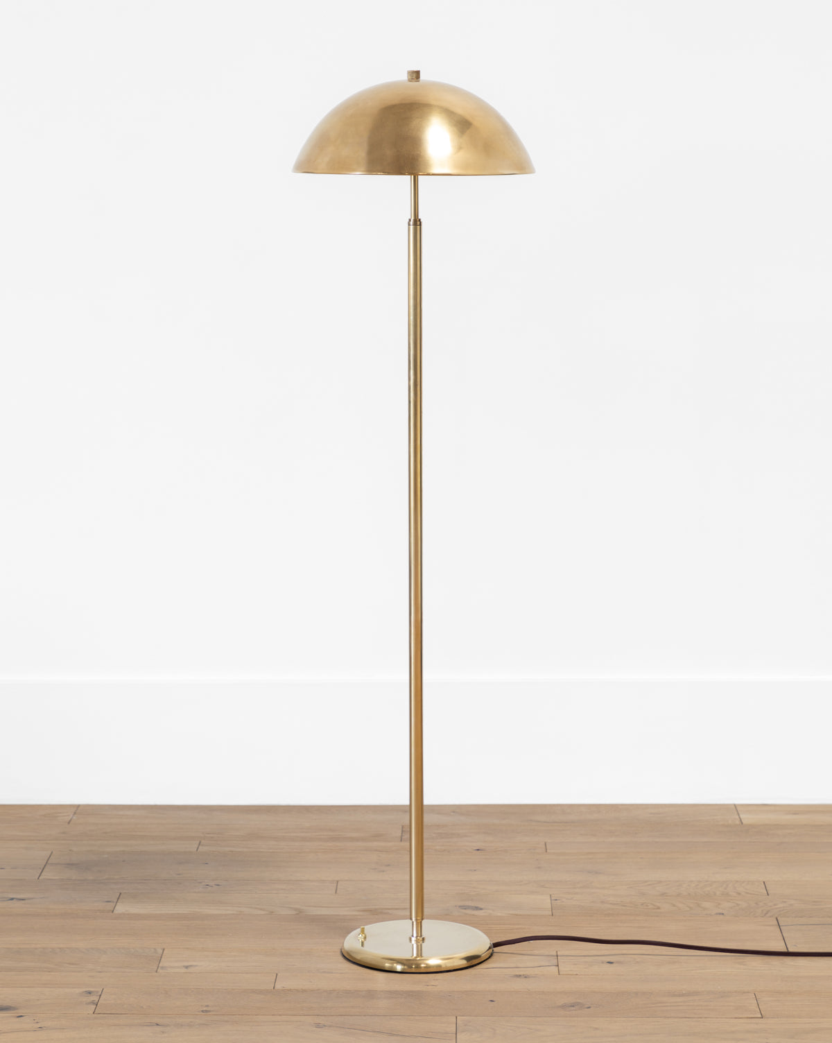 The Makerspalm Aldorno Floor Lamp features a slim pole, wide round shade, and round base on a light wooden floor against a white wall, with a black power cord extending from its base. Vendixs