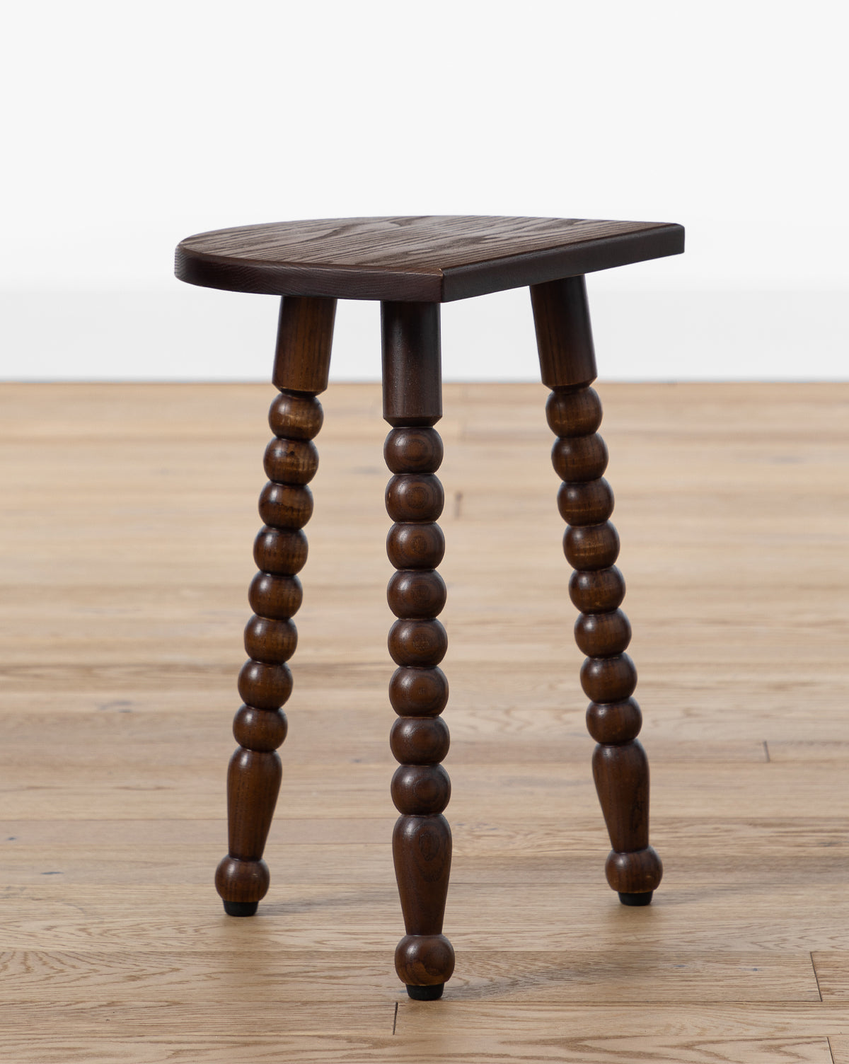 The Alice Wood Stool features a compact, vintage-inspired design with a triangular ash wood seat and turned legs, shown on a light wooden floor against a plain white background, by Vendixs
