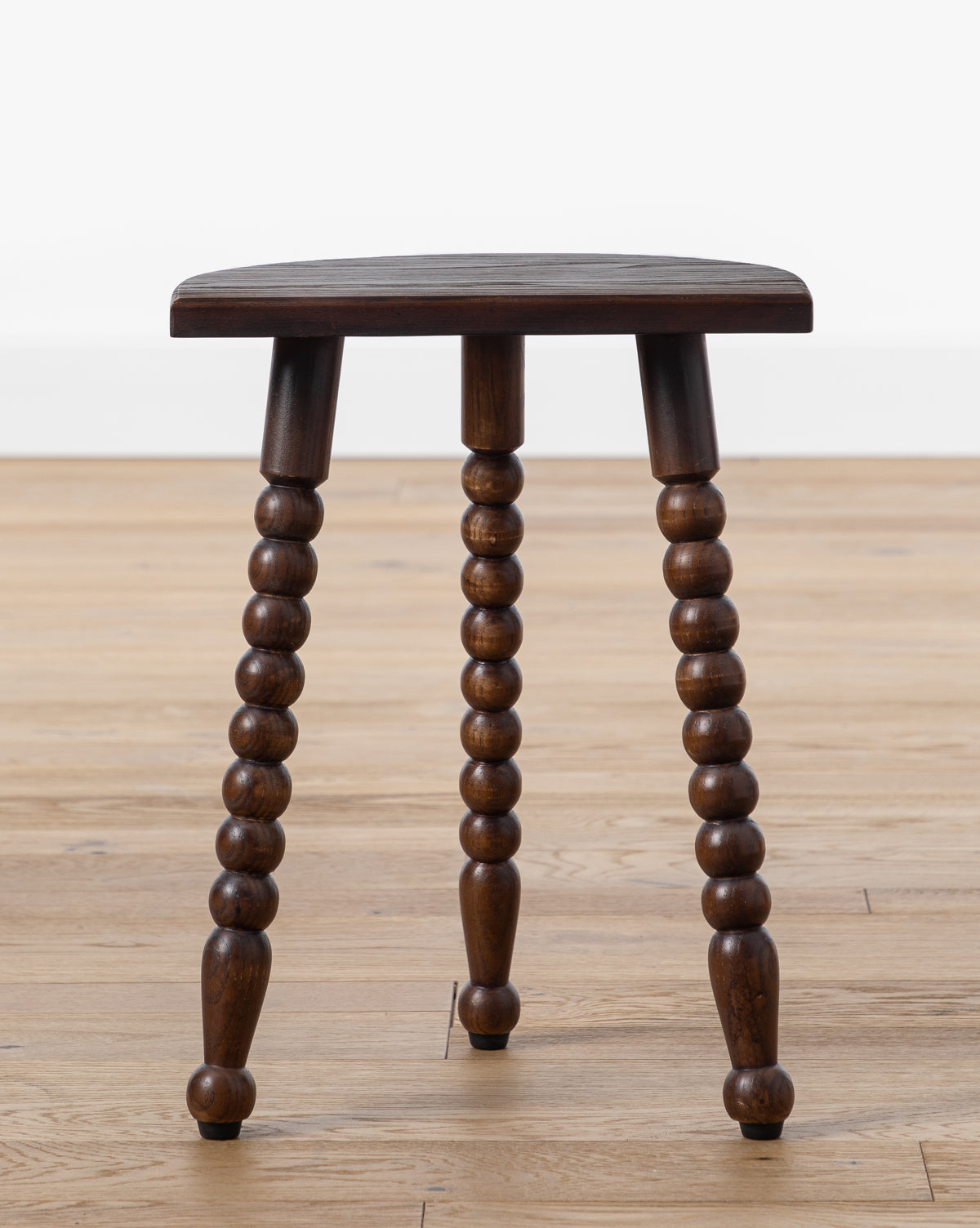 The Alice Wood Stool by Makerspalm is a vintage-inspired ash wood stool with a dark polished seat and turned, beaded legs, shown on a light wood floor against a plain white background by Vendixs