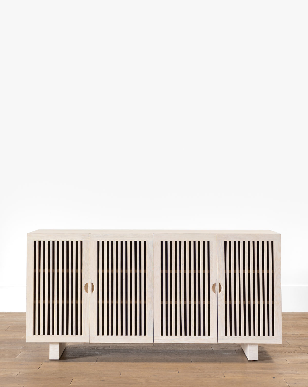 The Makerspalm Alvin Sideboard is a modern solid oak piece with four doors and vertical slats, standing on low square legs, shown here on a light wood floor beside a plain white wall. Vendixs