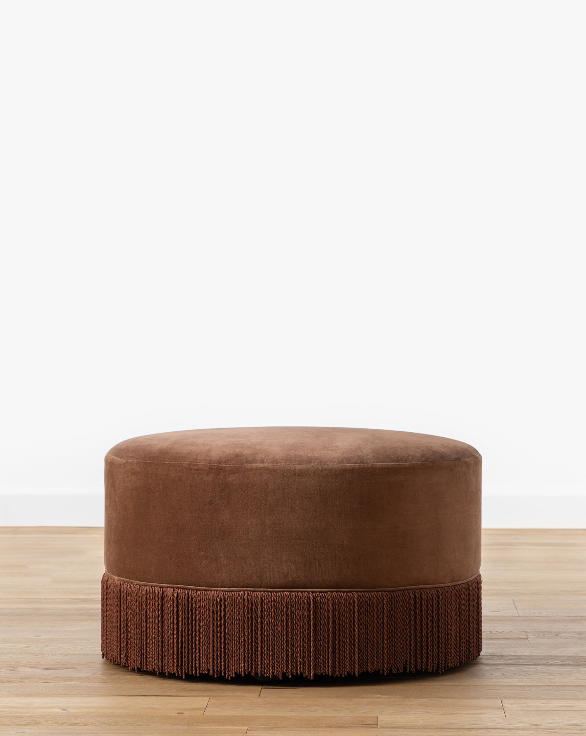 Certainly! Here’s your blended sentence with Vendixs at the end: The Makerspalm Astrid Fringe Ottoman, a round brown velvet piece with mid-century design and decorative fringe trim, sits on a light wood floor against a plain white wall, Vendixs