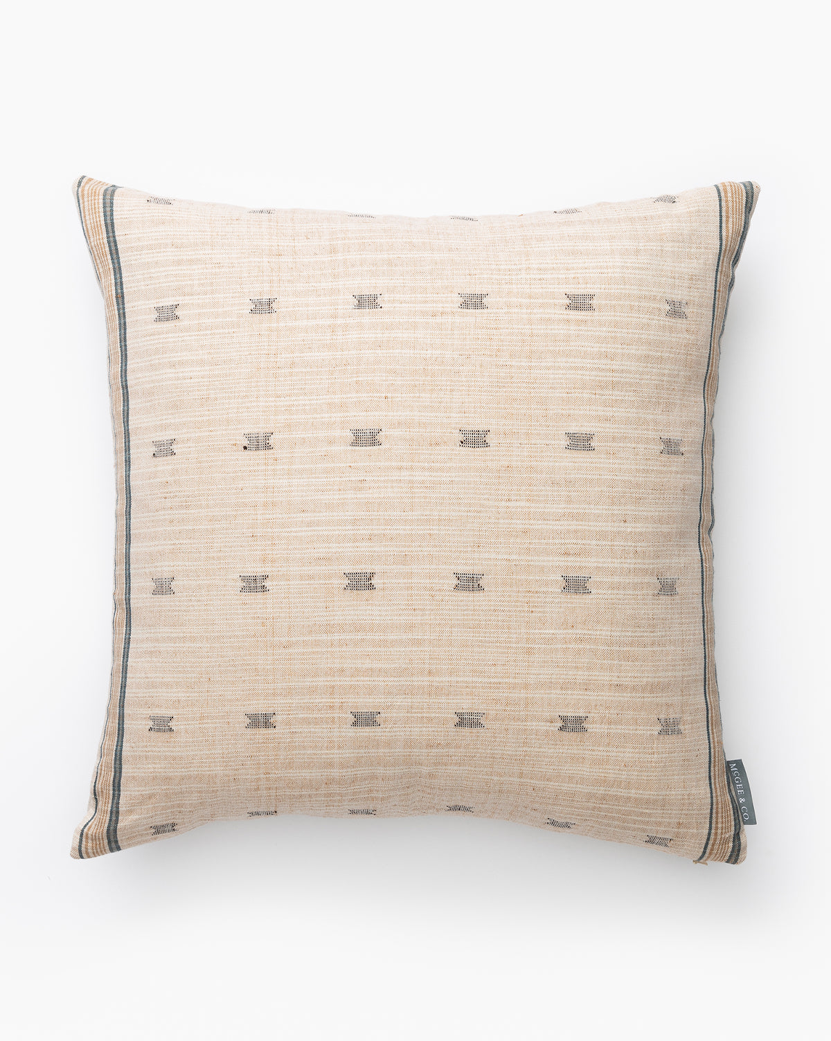 The Bali Pillow Cover is a square beige cotton pillow cover with subtle horizontal lines, small dark geometric patterns, thin dark borders at the edges, and a small fabric tag on one corner, shown against a white background by McGee & Co.