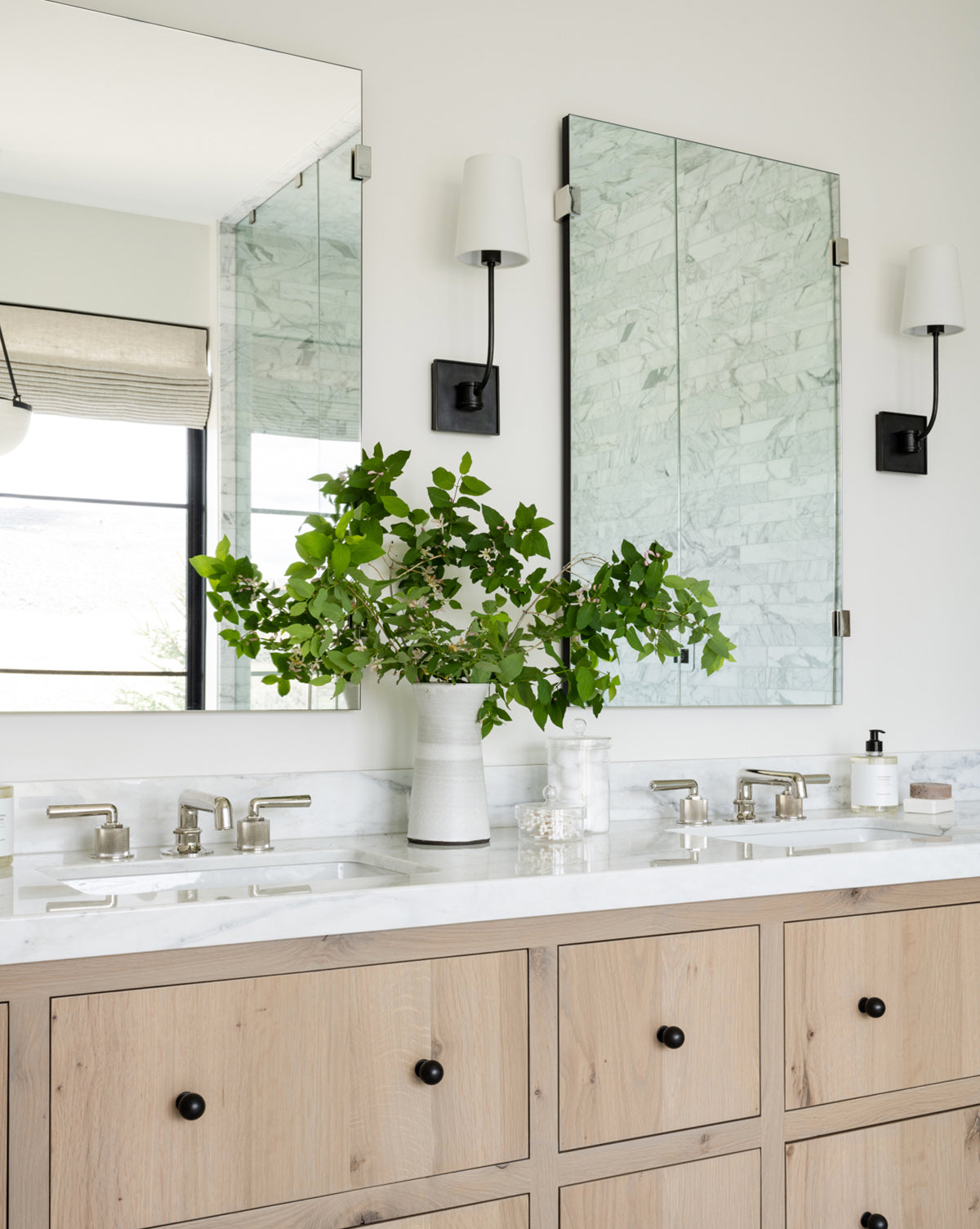 Modern bathroom featuring a light wood double vanity, white marble countertop, two Ivana Mirrors, burnished brass wall sconces, a vase with green branches, and window reflections with a roman shade, by Vendixs